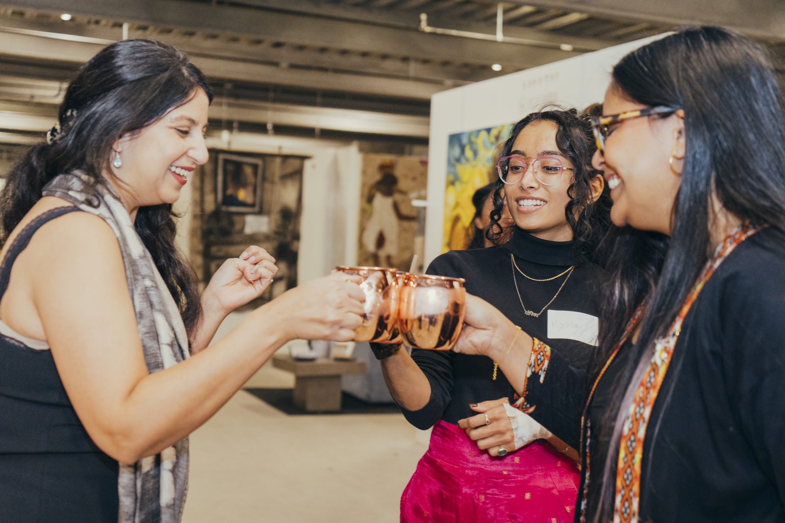 Three women raising copper mugs at the Brown Girl Angels Investing in Art evening — a toast among didis.